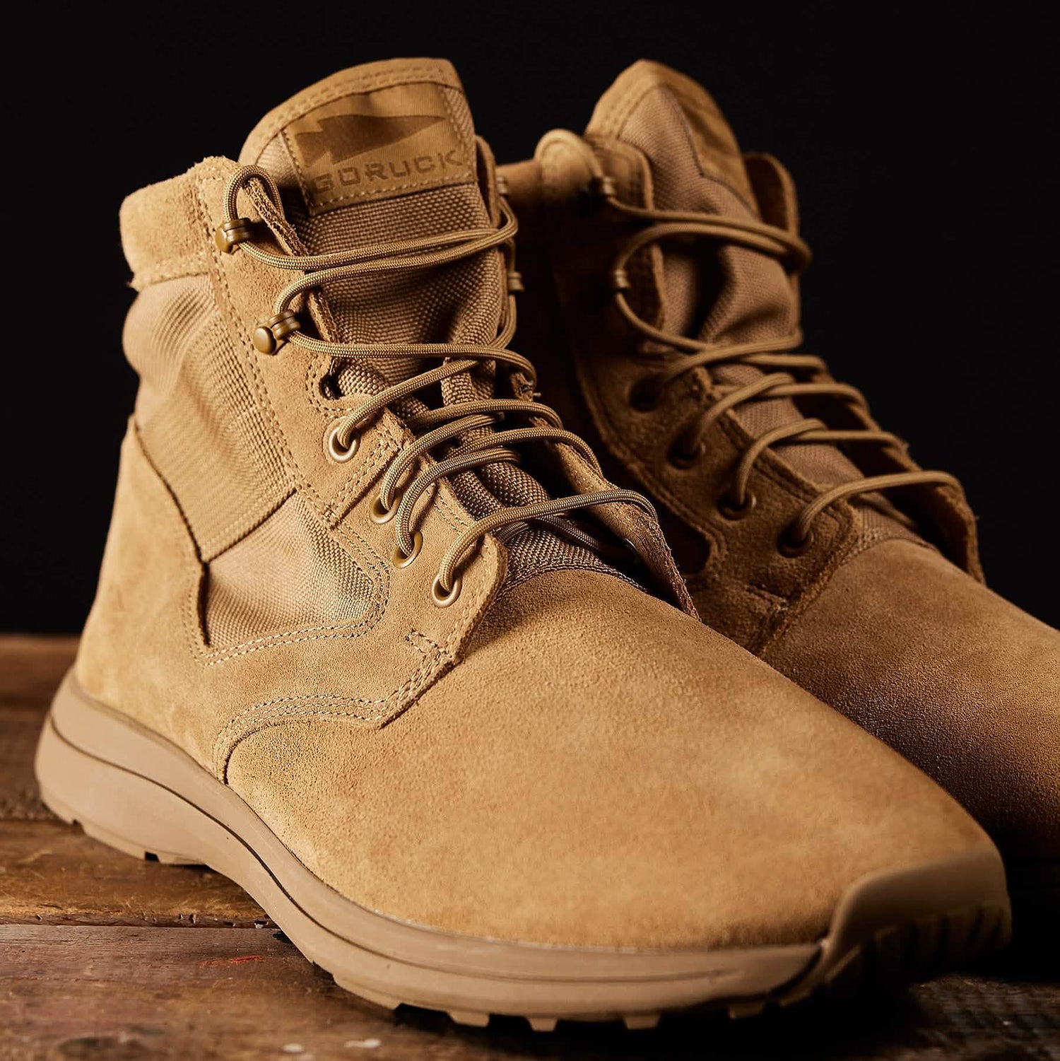 A pair of tan-colored GORUCK MACV-1 Hi-Speed - Mid Top boots, renowned for their lightweight design, rest on a wooden surface against a dark backdrop. These boots feature thick soles, durable laces, rugged stitching, and a brand patch on the tongue, highlighting their suitability for rucking adventures.