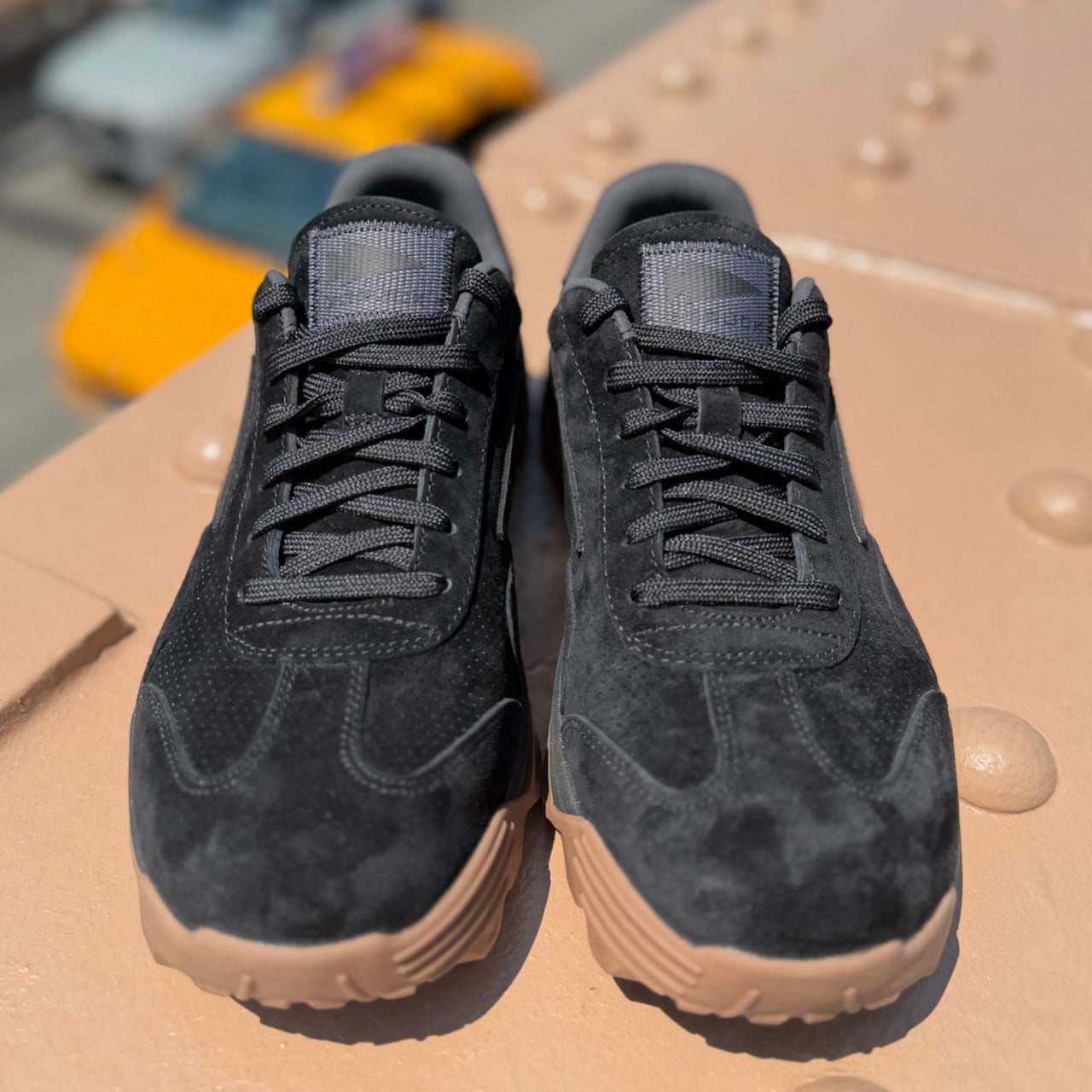 A pair of Mackall 45 Black + Gum sneakers with black suede uppers and tan soles are placed on a textured beige surface outdoors.