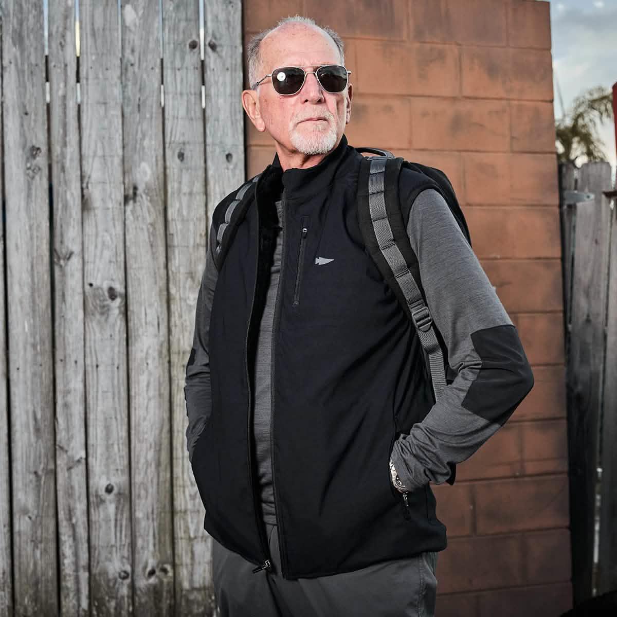 Older man wearing sunglasses, black softshell vest and grey long sleeve shirt with backpack against wooden and brick fence