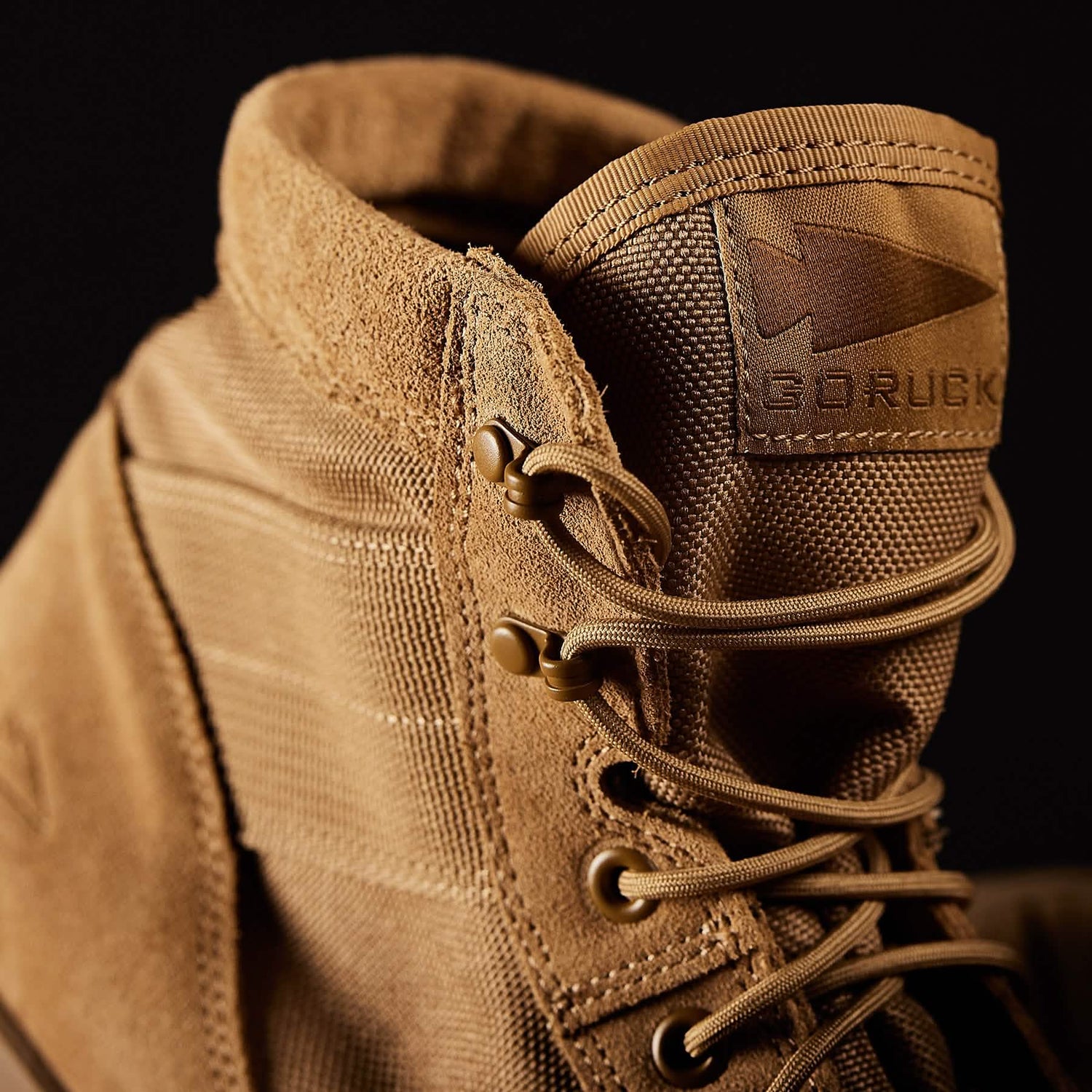 The MACV-1 Hi-Speed - Mid Top by GORUCK is shown in a close-up, revealing its rugged brown construction adorned with detailed stitching and sturdy laces. The durable material, ideal for outdoor adventures, is highlighted by a small arrowhead logo on the tongue that identifies it as a MACV-1™ rucking boot. Set against a dark background, the lightweight boot prominently stands out.