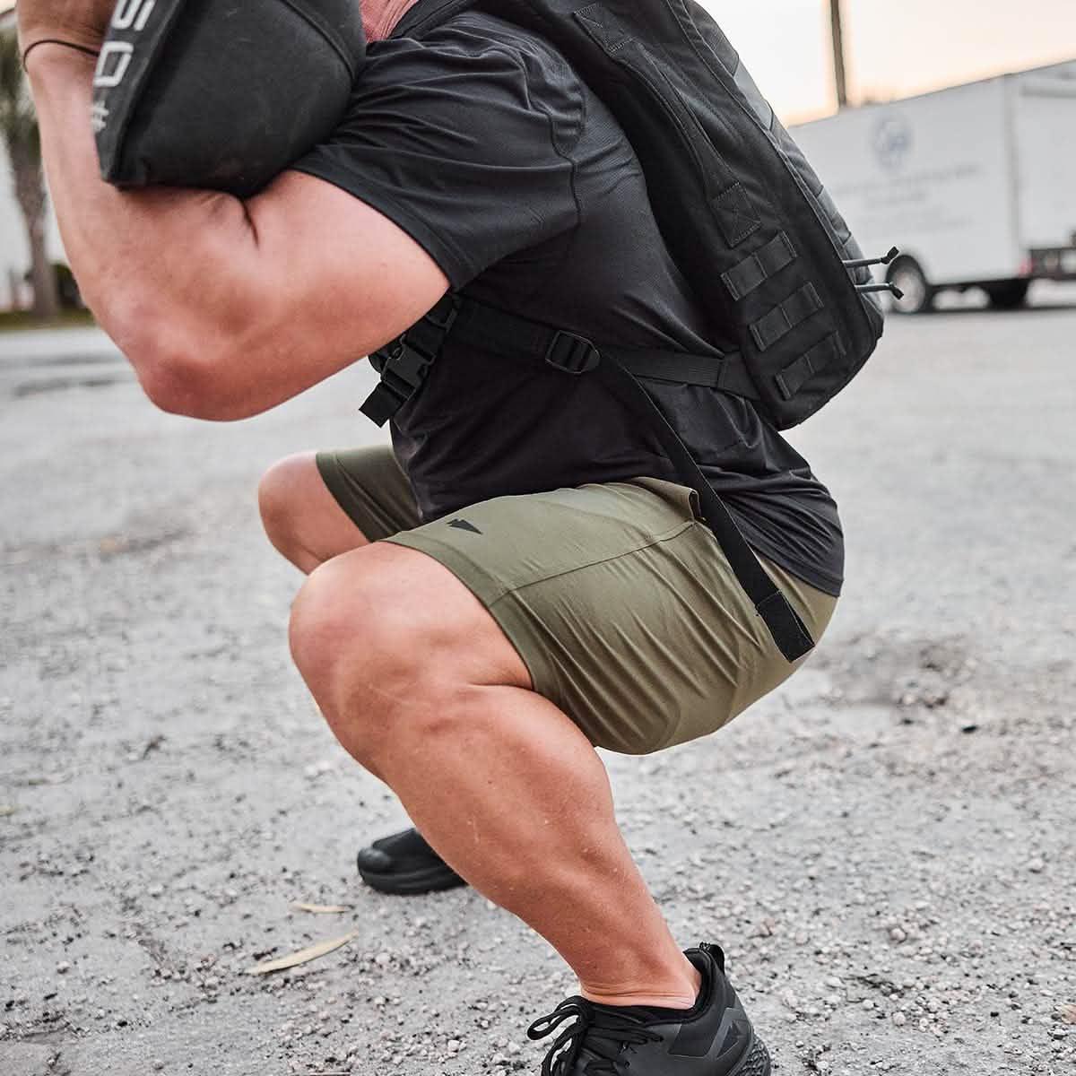 Man squatting outdoors in green GORUCK shorts, black shirt, and rucksack during rucking workout
