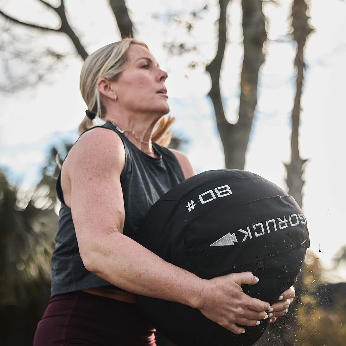 Woman exercising outdoors holding a black GORUCK 80-pound weighted sandbag during fitness training