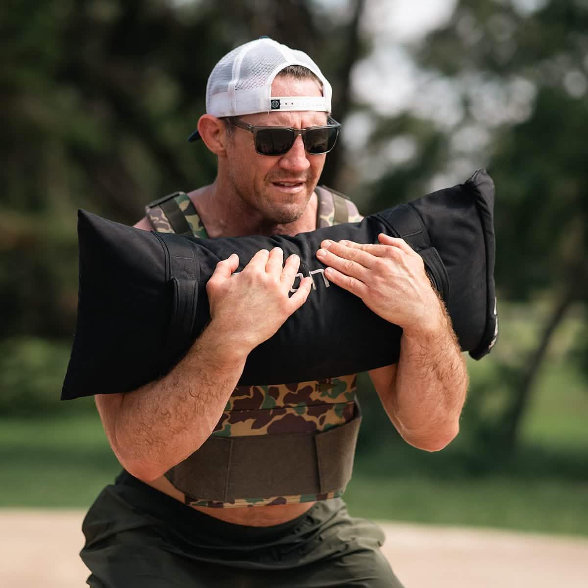 Man outdoors wearing GORUCK camo vest, holding weighted sandbag during rucking workout