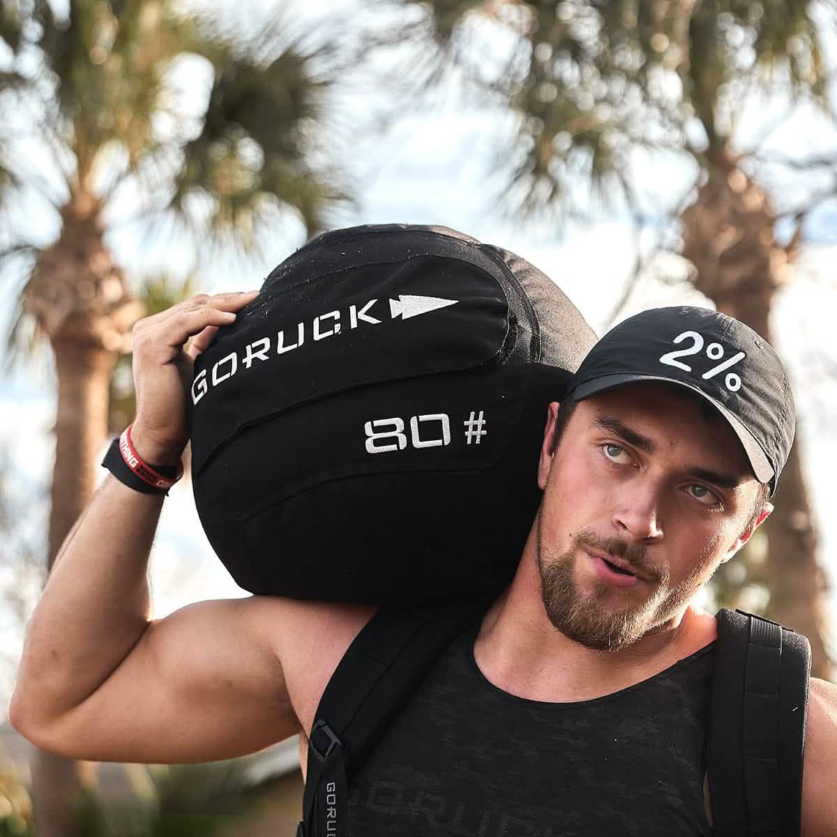 Man carrying a black 80-pound GORUCK weighted bag on his shoulder outdoors with palm trees in the background