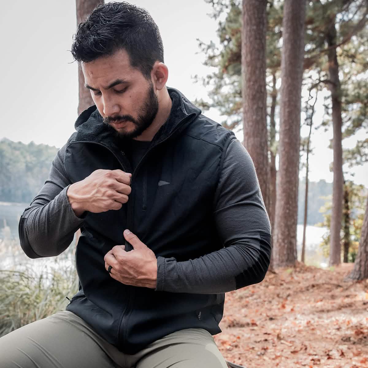 A man sits outdoors by a tranquil lake, embraced by nature's greenery. Dressed in the GORUCK Vest of Power made from ToughDry® + Fleece over a long-sleeve gray shirt and green pants, he thoughtfully adjusts the zipper. The serene, forested backdrop mirrors his eco-friendly mindset.
