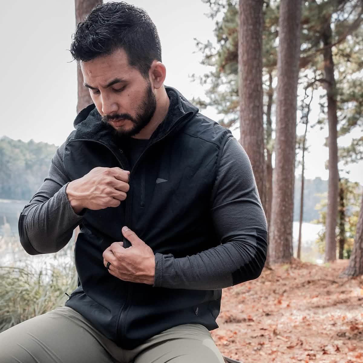Man outdoors zipping black GORUCK vest over gray long-sleeve shirt in forest setting with pine trees