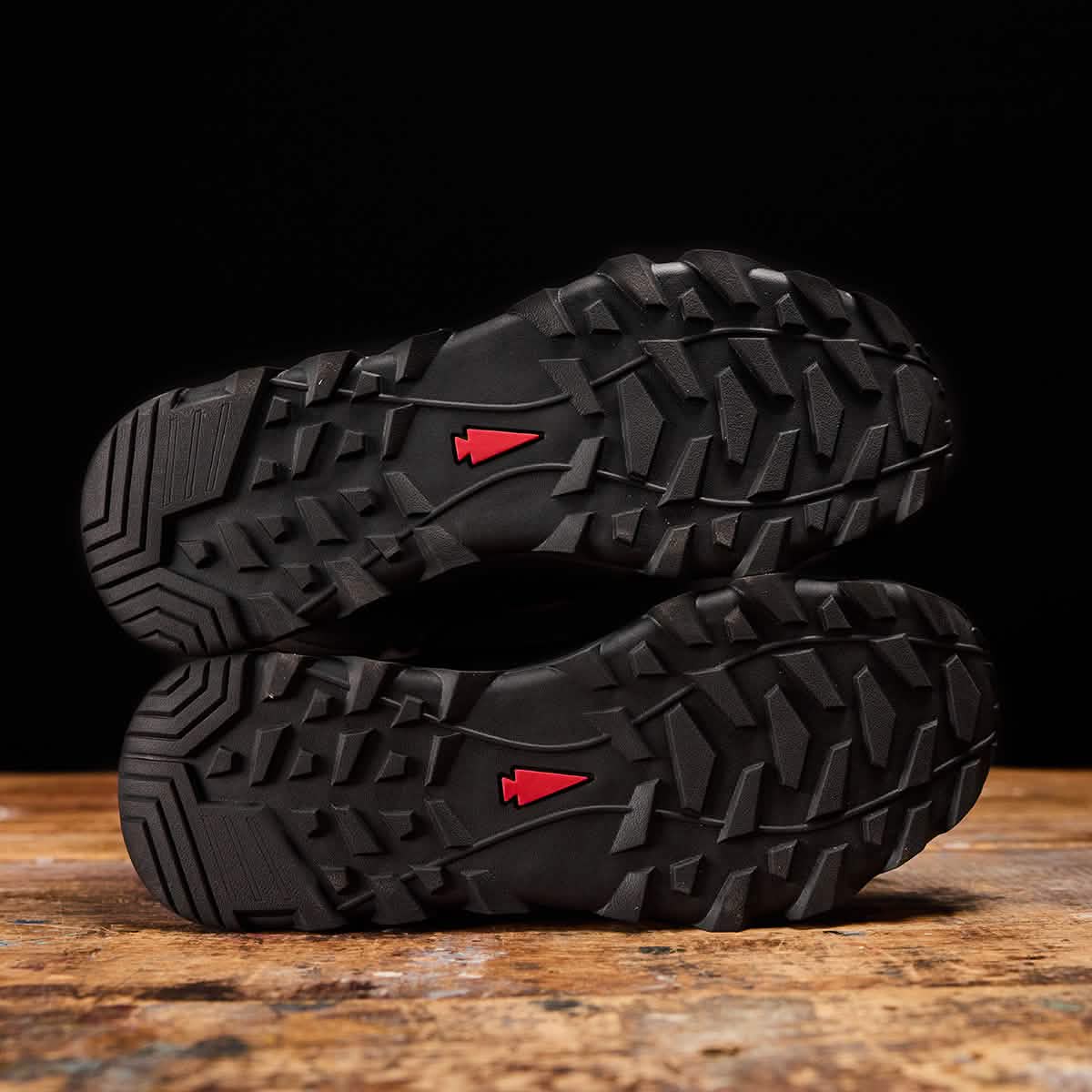 The image showcases the soles of the MACV-2 Safety Boot - High Top by GORUCK, positioned sole to sole. The tread pattern, resembling Special Forces specifications, features a robust design with a small red arrow at the center of each sole, all against a dark background.