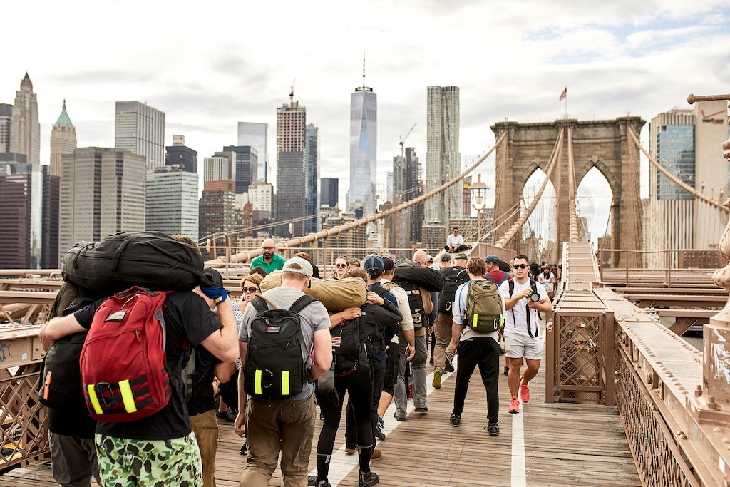 A group of people with backpacks walking across the Brooklyn Bridge with the Manhattan skyline in the background.