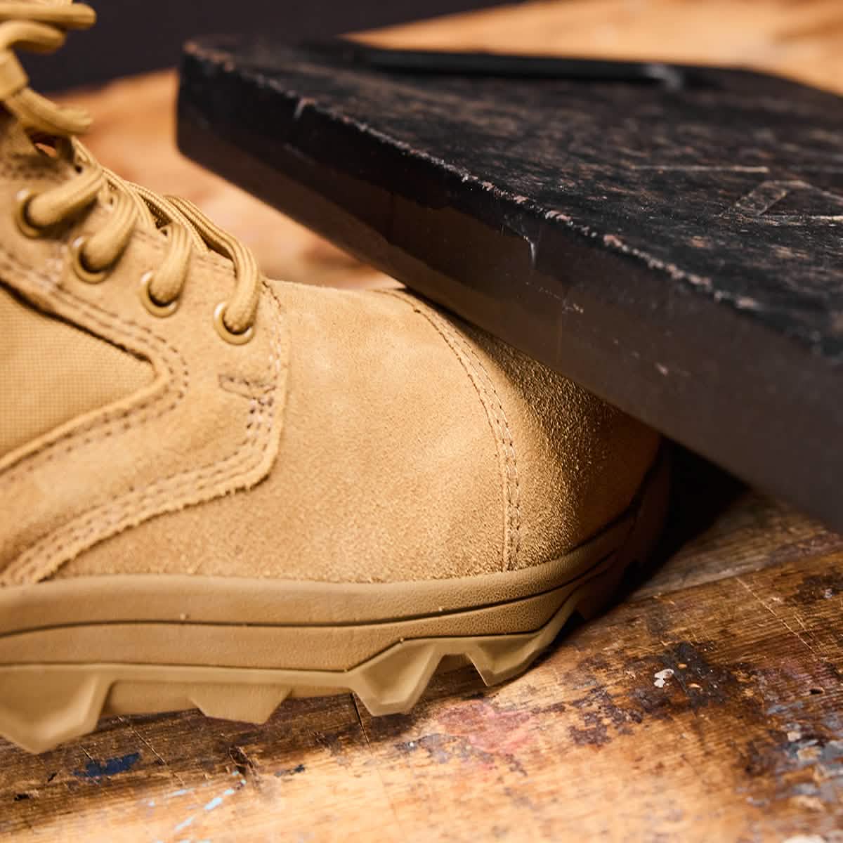 The MACV-2 Safety Boot - Mid Top by GORUCK is displayed in close-up, showcasing its tan color and rugged soles that meet Special Forces standards by shielding a foot from a heavy black object pressing down. The boot is positioned on a worn, wooden surface.