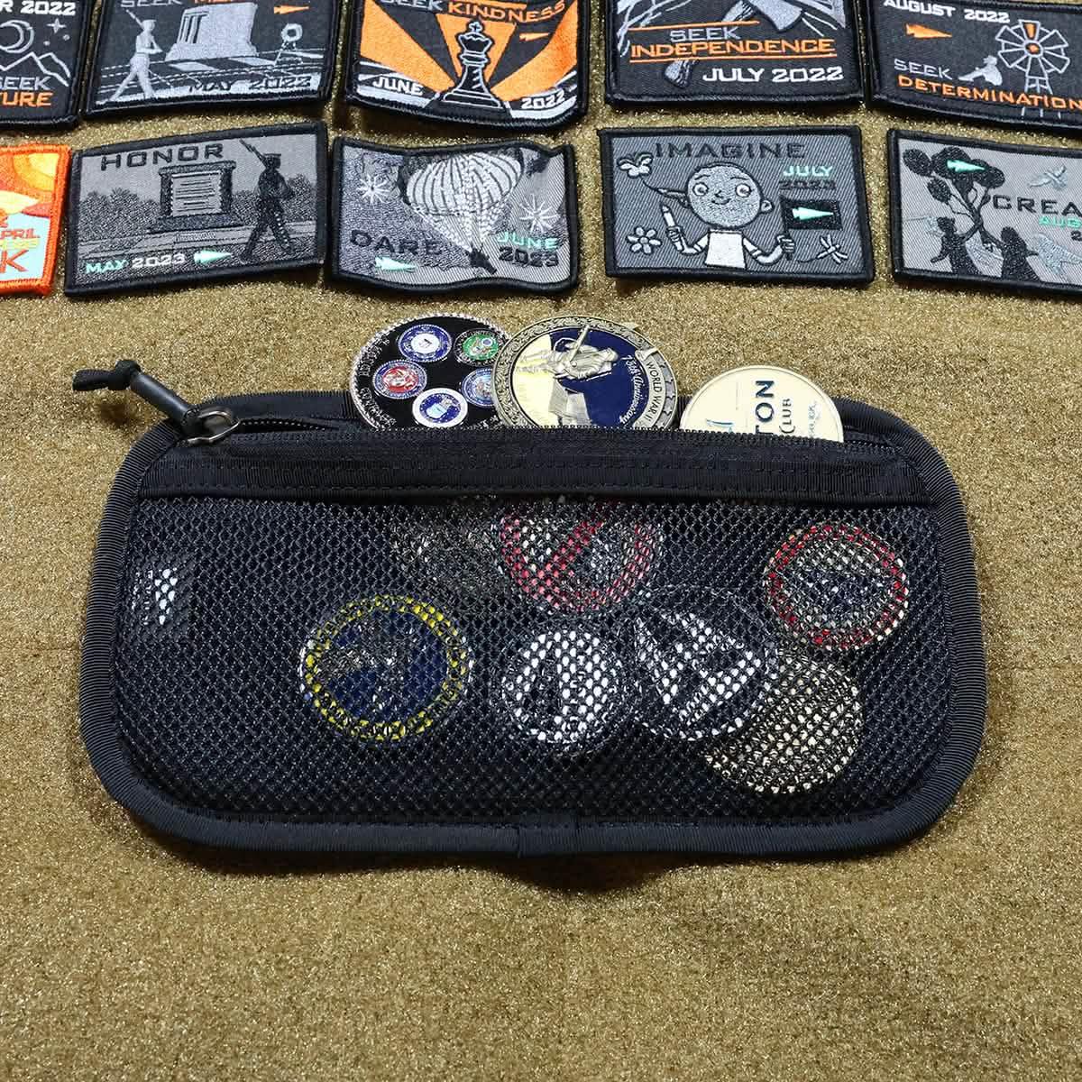 Black mesh zippered pouch partially open showing GORUCK challenge coins and rucking event patches on tan surface