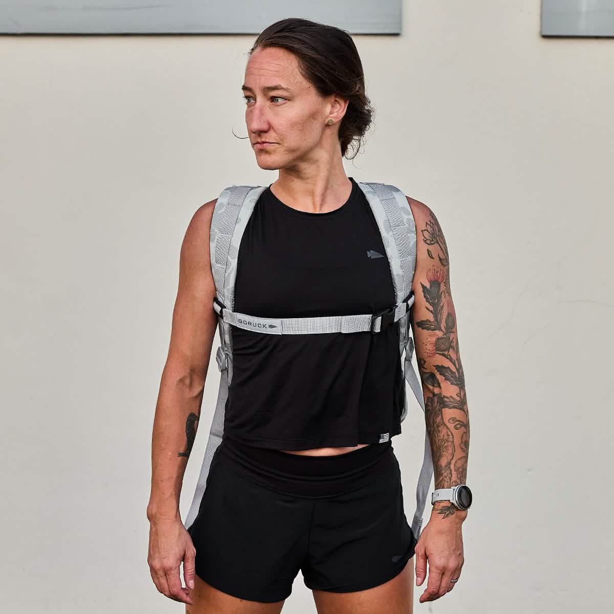 Athletic woman with tattoos in a black outfit stands against a plain light wall, looking sideways, wearing the GR0 - Ripstop ROBIC® minimalist everyday carry backpack.