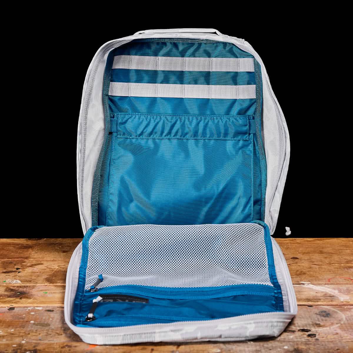 Open GR0 - Ripstop ROBIC® compact rucksack in gray and blue on a wooden surface, revealing multiple compartments and mesh pockets inside—ideal for minimalist everyday carry.