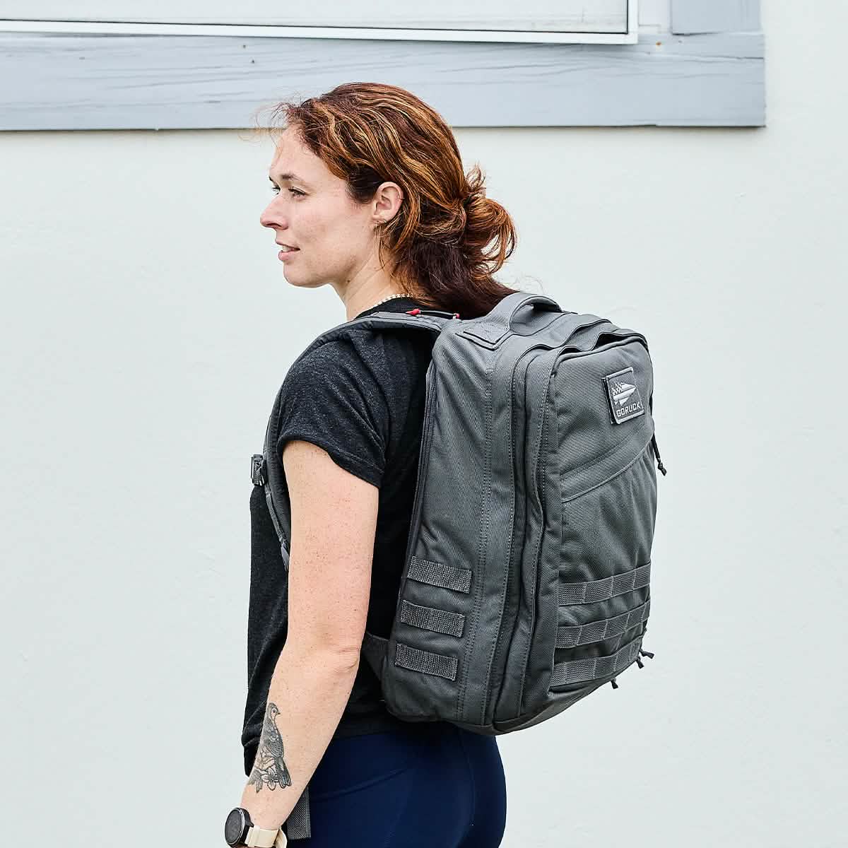 A person with reddish-brown hair stands sideways, carrying a large gray GR2 - Cordura ruck by GORUCK. They wear a black T-shirt and have a tattoo on their left forearm, set against a plain light-colored wall, exuding an air of understated strength reminiscent of Special Forces precision.