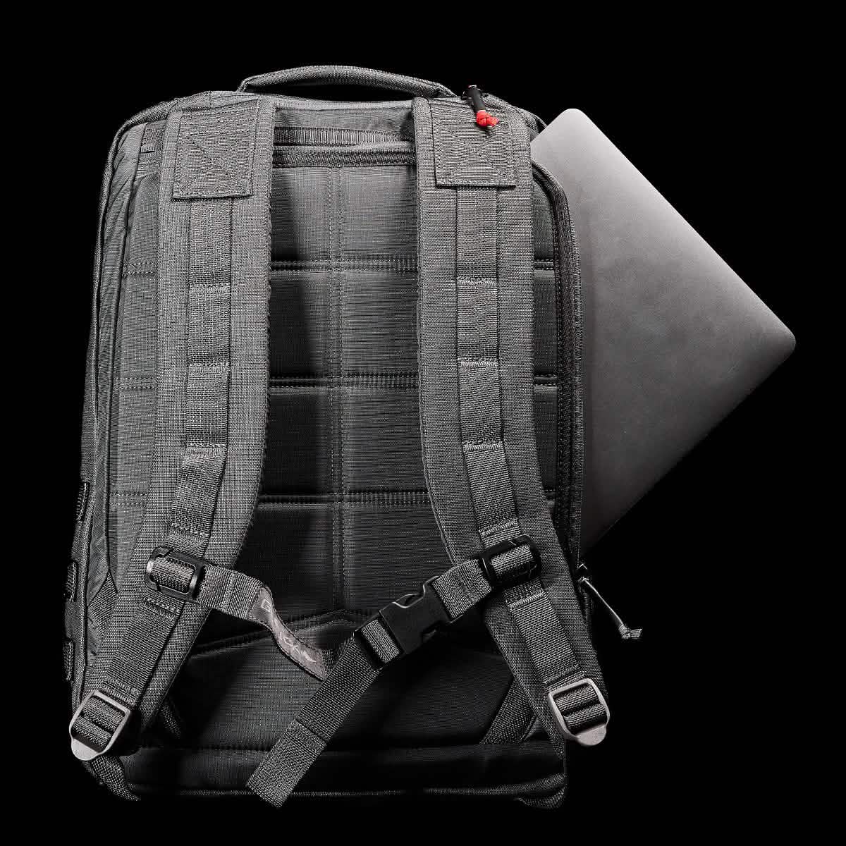 A black GORUCK GR2 backpack with padded shoulder straps and a quilted back sits against a black background. A laptop is partially visible, either being inserted or removed from the side pocket. The scene is well-lit, emphasizing the backpack's texture, details, and its Special Forces quality design.