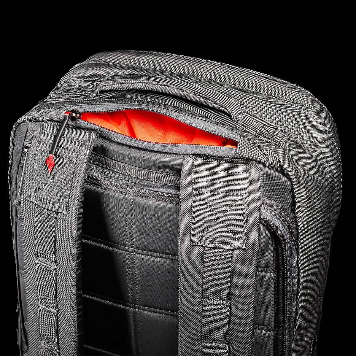 A close-up of a black GORUCK GR2 - Cordura backpack shows an open zipper revealing its orange interior. Inspired by Special Forces design, this backpack features padded shoulder straps and a grid pattern on the back, with zippers that have small red tab accents.