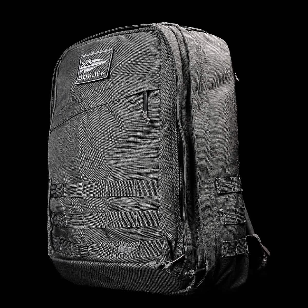 A rugged GR2 backpack, in dark gray against a black background, exemplifies Special Forces quality. It has a zippered front pocket, prominently displays the GORUCK logo patch, and includes multiple webbing strips for attachments. The backpack provides a sturdy and durable appearance, making it ideal for carry-on compliant travel.