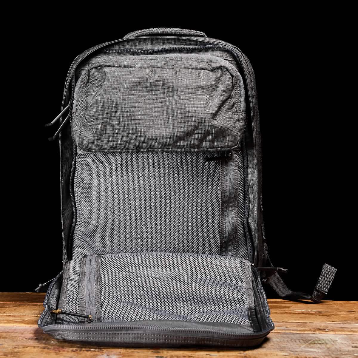 The GORUCK GR2 backpack, featuring multiple compartments such as a front pocket and a zippered section, is showcased in gray against a black background on a wooden surface. Designed to meet Special Forces quality standards, it offers excellent durability and carry-on compliant convenience for those who seek reliability.