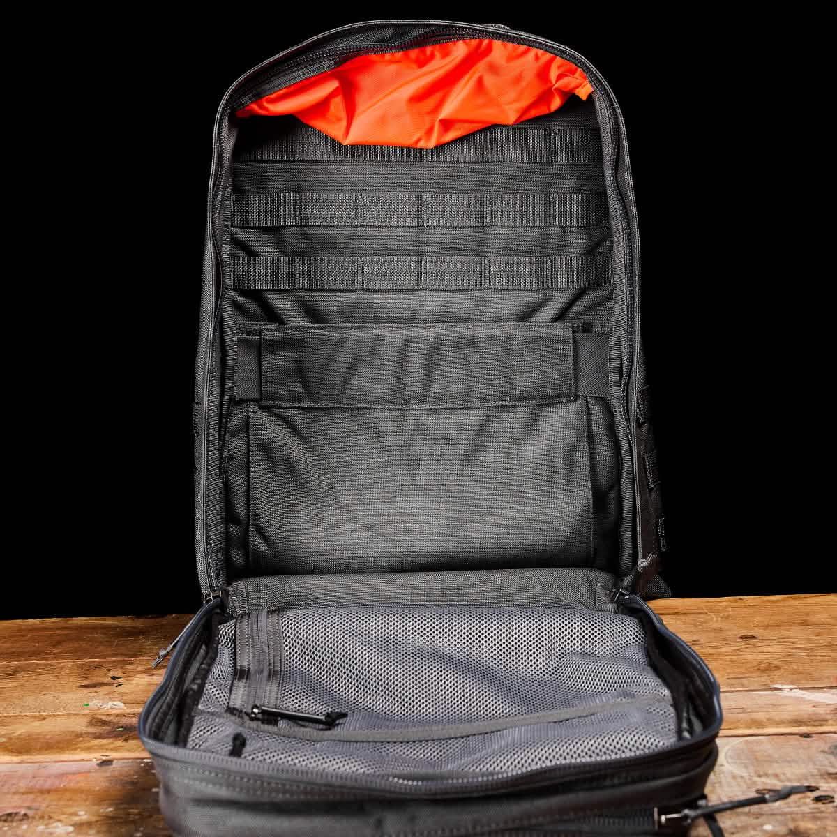 The GR2 - Cordura by GORUCK, an open black backpack, reminiscent of those used by Special Forces, sits on a wooden table, showcasing its spacious interior. With an orange lining, multiple compartments, and a mesh pocket for organizing essentials, it highlights its impressive storage capacity against a plain black background.
