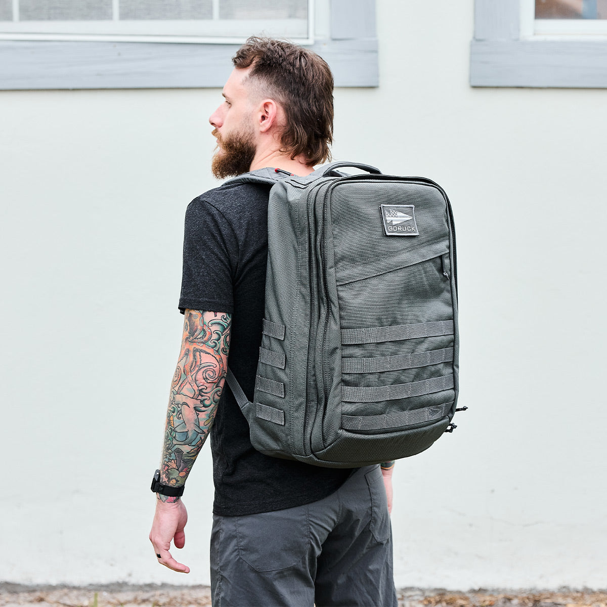 Man with tattooed arm wearing a black t-shirt and gray pants carrying a large GORUCK grey tactical rucksack outdoors