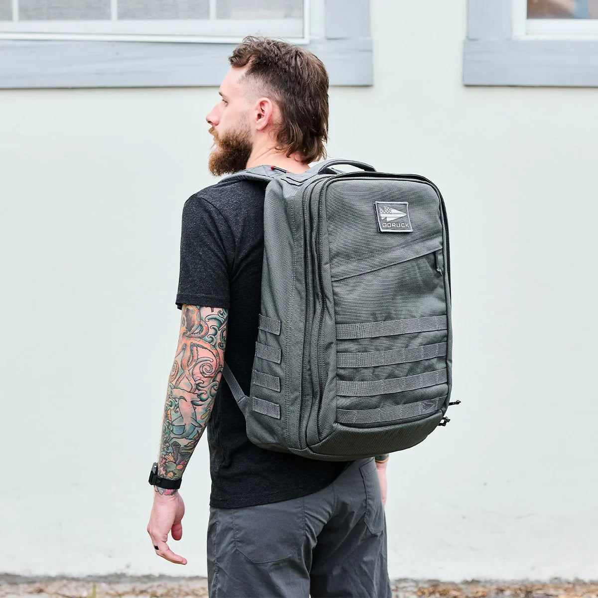 Man with tattooed arm wearing a black t-shirt and gray pants carrying a large GORUCK grey tactical rucksack outdoors
