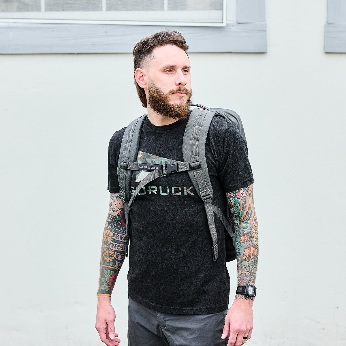 Man with beard and tattoos wearing black GORUCK t-shirt and gray rucking backpack against light wall