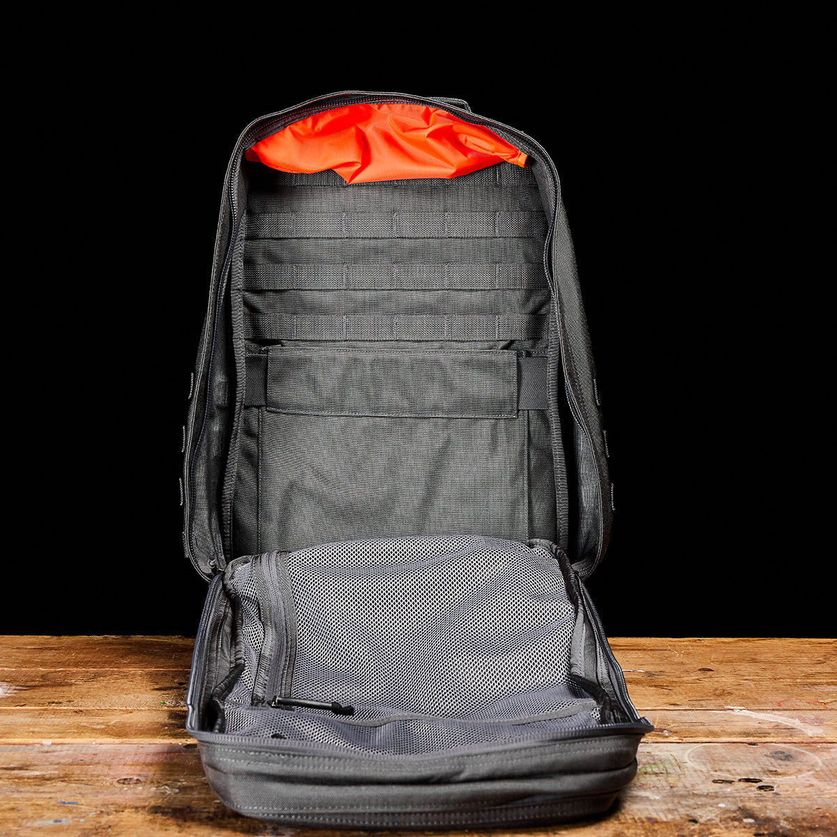 Open GR2 - Ballistic Nylon Cordura rucksack in gray with orange lining, empty and standing upright on a wooden surface against a black background.