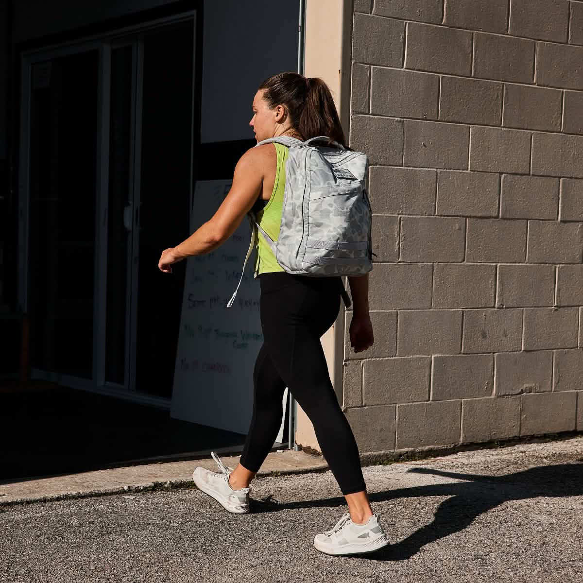 A woman in workout clothes carries the GR0 - Ripstop ROBIC® rucksack as she walks outdoors along a building wall in bright sunlight.