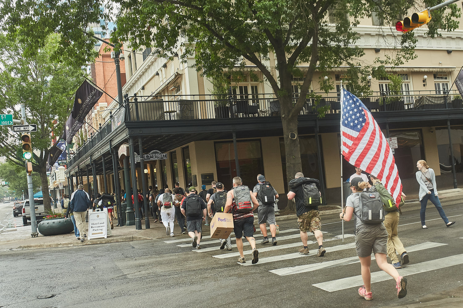 A group of people with backpacks cross a city street, one carrying a large American flag.