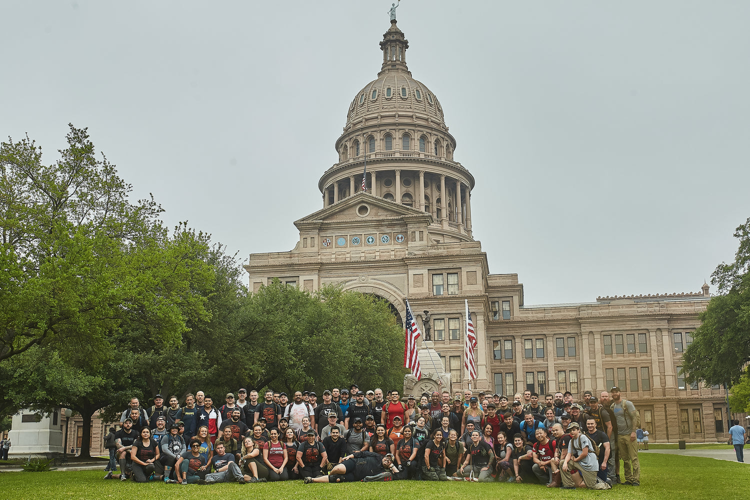 A large group poses on the lawn in front of the Texas State Capitol building on a cloudy day.