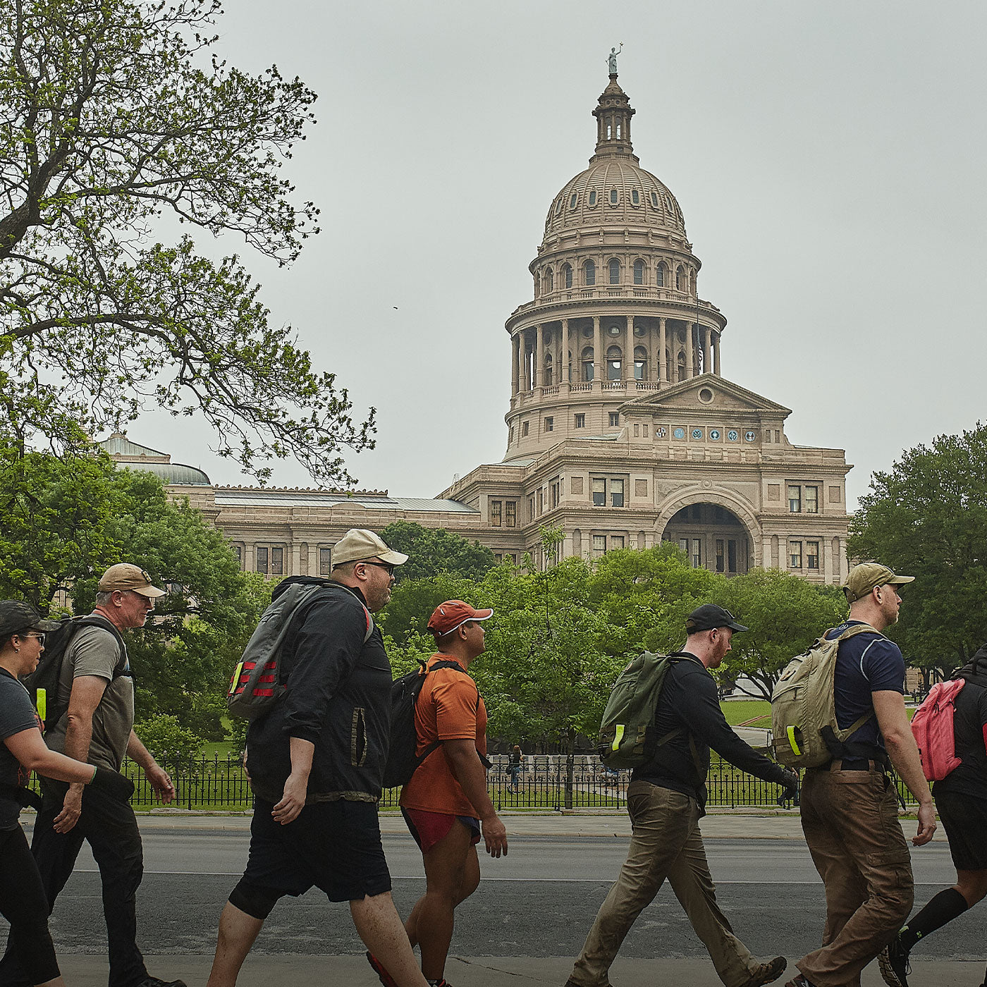 A group of people with backpacks walk past a large domed capitol building on a cloudy day.