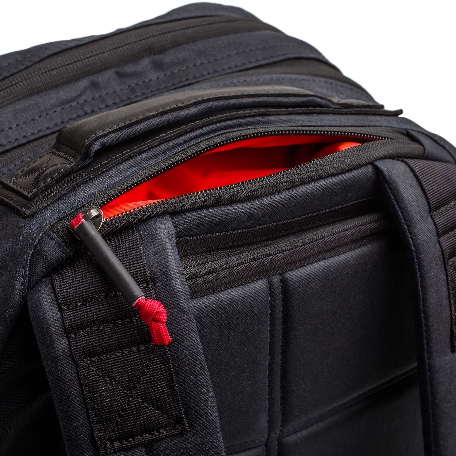 Close-up of the Double Compartment Bullet Ruck - Heritage Waxed Canvas - 18L backpack in black, featuring a red zipper pull and a bright orange interior lining.