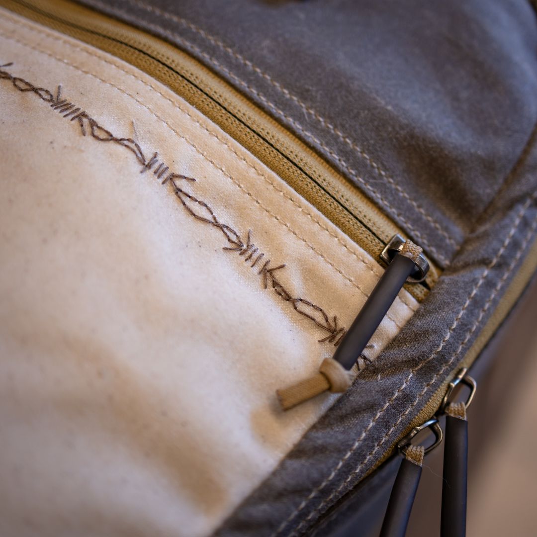 Close-up of a limited edition gray and cream-colored backpack featuring a zippered pocket with handcrafted barbed wire embroidery along the seam.