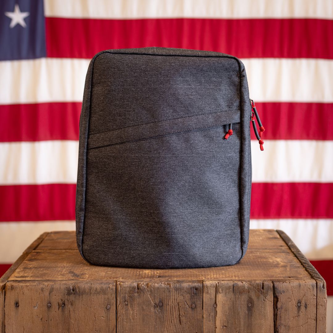 A handmade, dark gray zippered backpack sits on a wooden crate in front of an American flag.