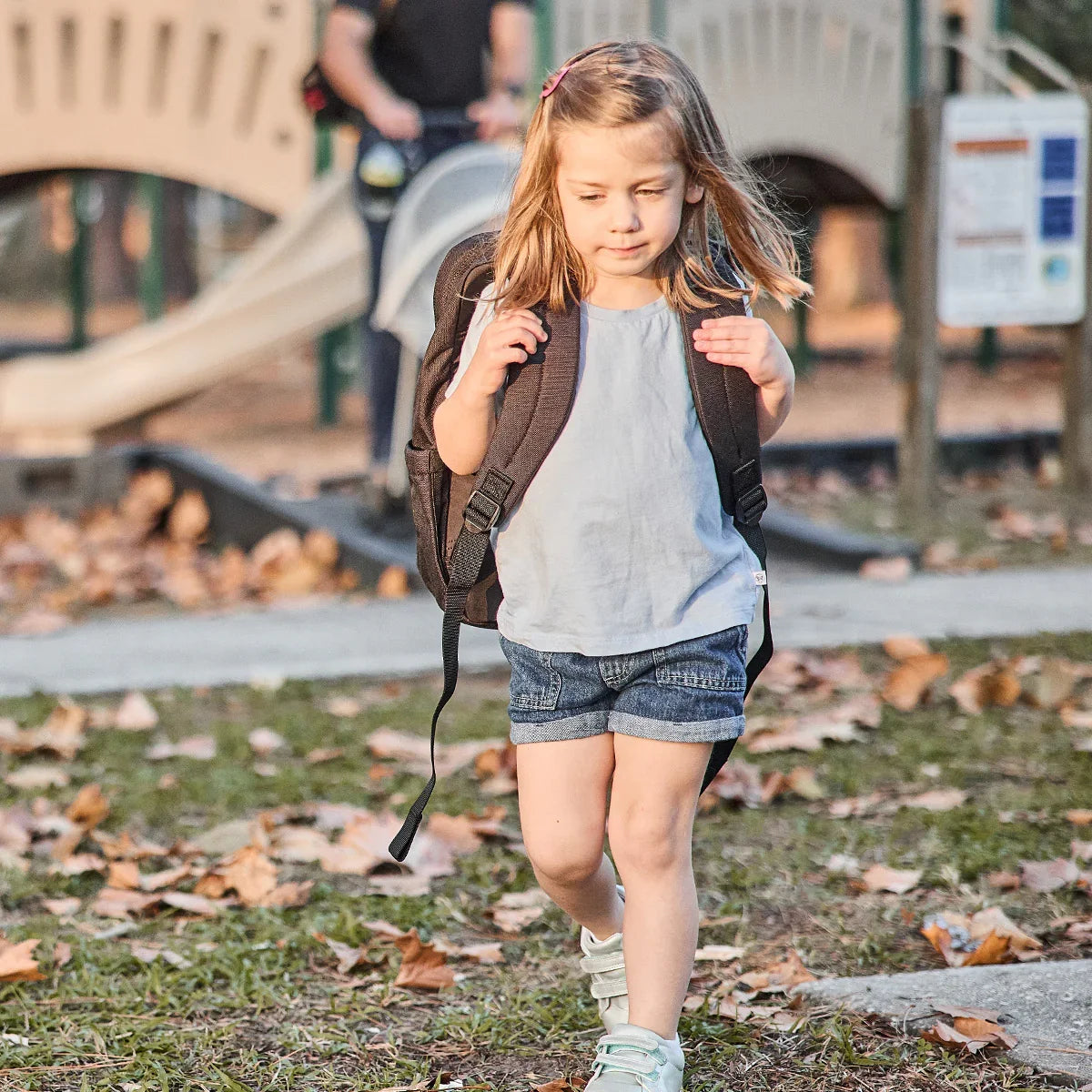Young girl wearing a GORUCK backpack outdoors near a playground, autumn leaves on ground