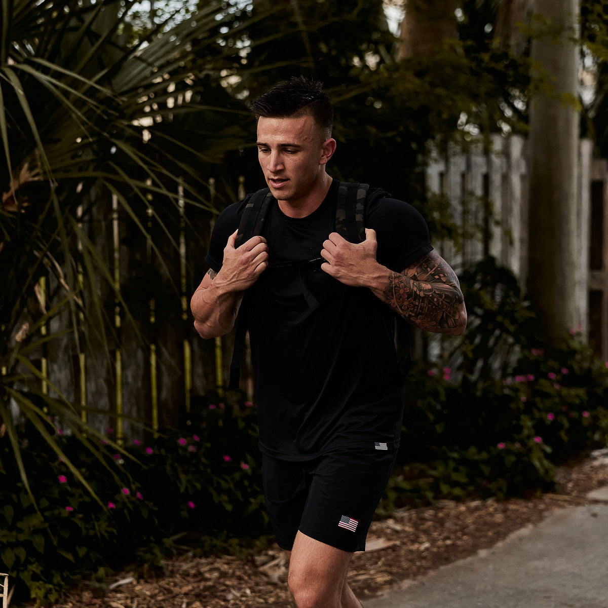 A tattooed man in black workout clothes carries a Ruck Plate Carrier 3.0, made from ballistic nylon, while walking outdoors.