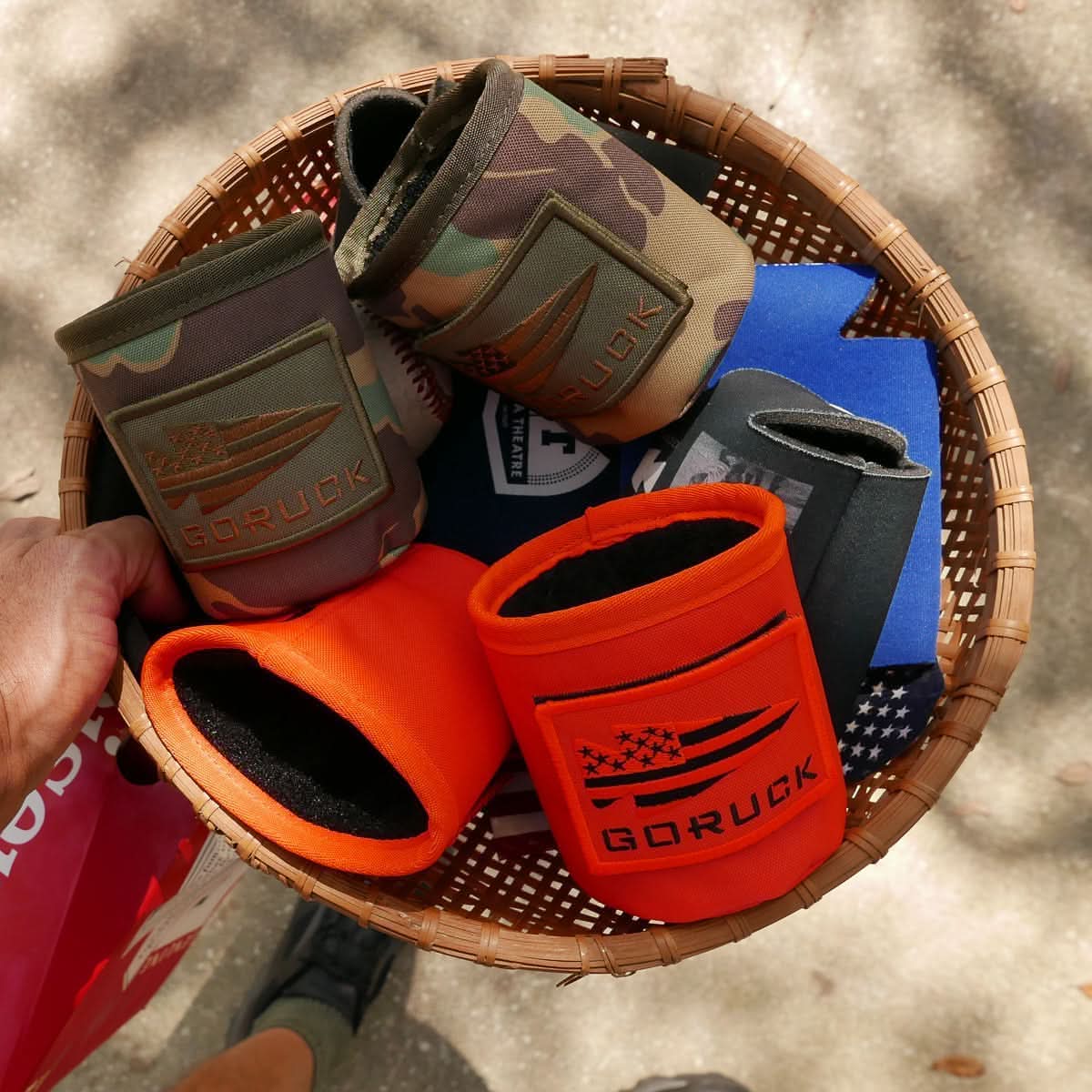 A hand holds a basket full of GORUCK Ballistic Beer Jackets made from 1000D CORDURA, featuring various colors and patterns like camo and orange. These durable can coolers are perfect for any BBQ or tailgate party.