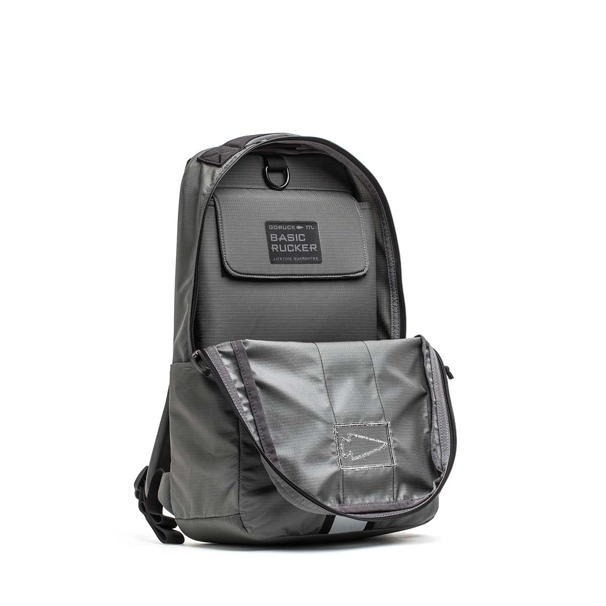 Gray Basic Rucker® backpack with front pocket partially open, revealing the inner compartment and a label reading