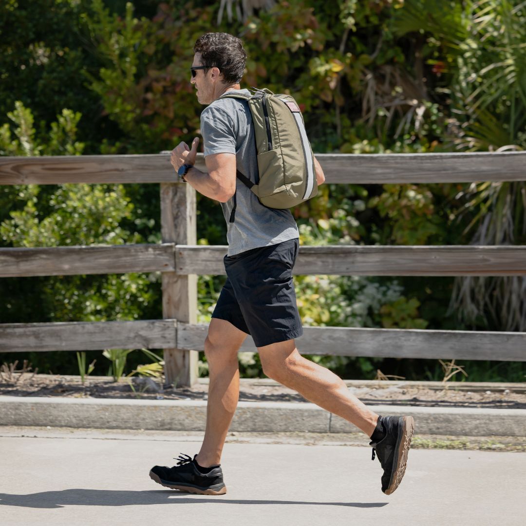 A man jogs on a sunny day, wearing sunglasses, a backpack, athletic clothes, and comfortable Merino Challenge Socks - Ankle (No-Show), with greenery in the background.