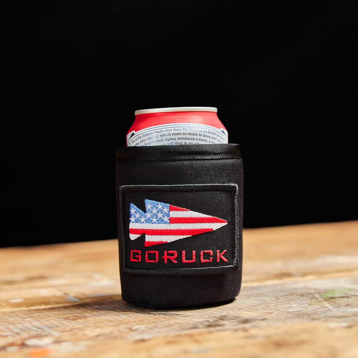 Red and white beer can inside a black GORUCK koozie with American flag arrow logo on wooden table