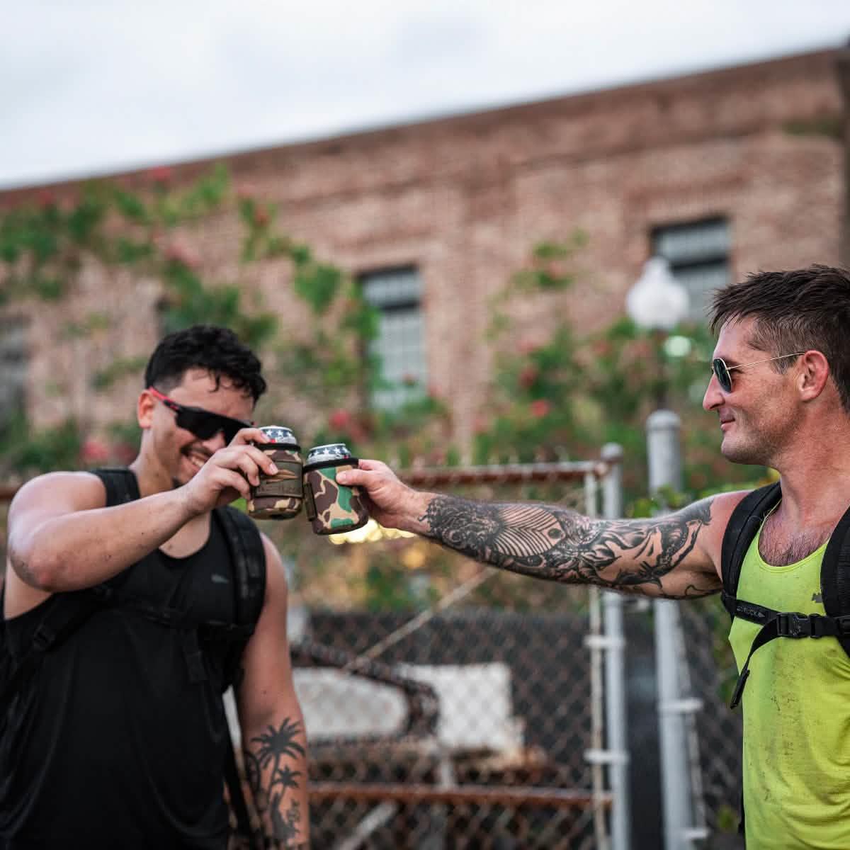 Two men wearing GORUCK gear clinking camo beer can coolers outdoors with a brick building background