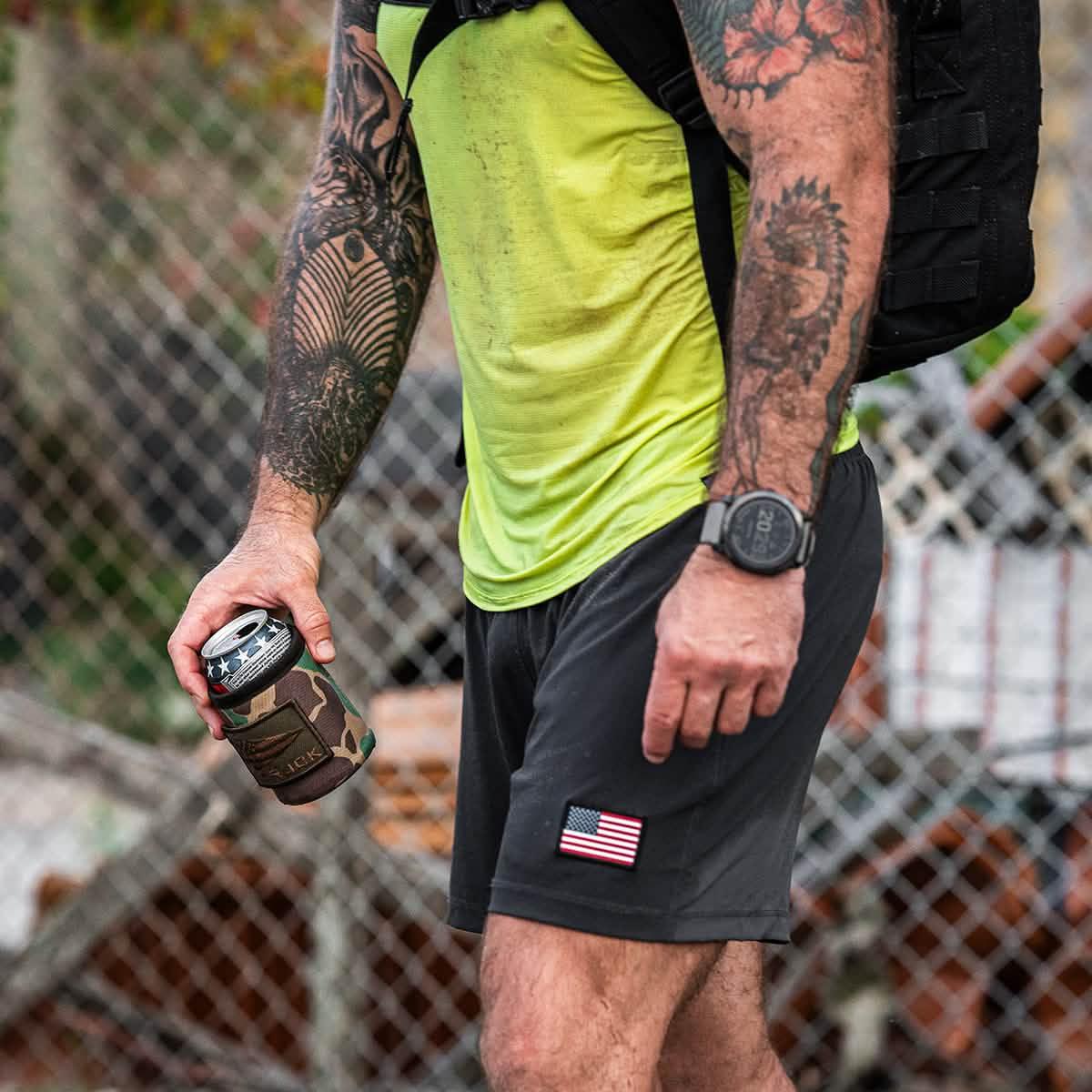 Man wearing bright yellow shirt and black shorts with American flag patch holding camo beer koozie outdoors