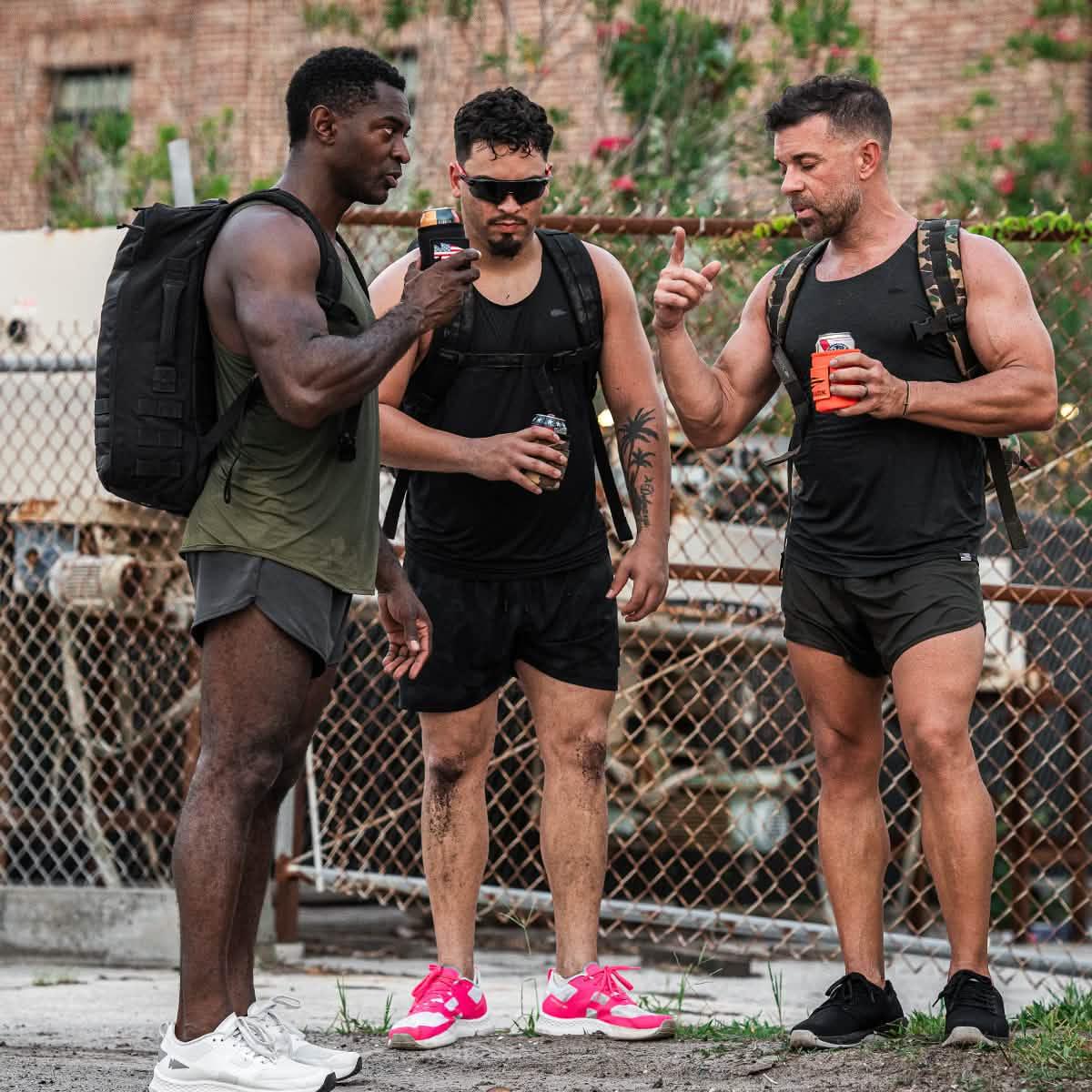 Three men in athletic gear and backpacks stand outside by a chain-link fence, holding drinks in GORUCK beer jackets.