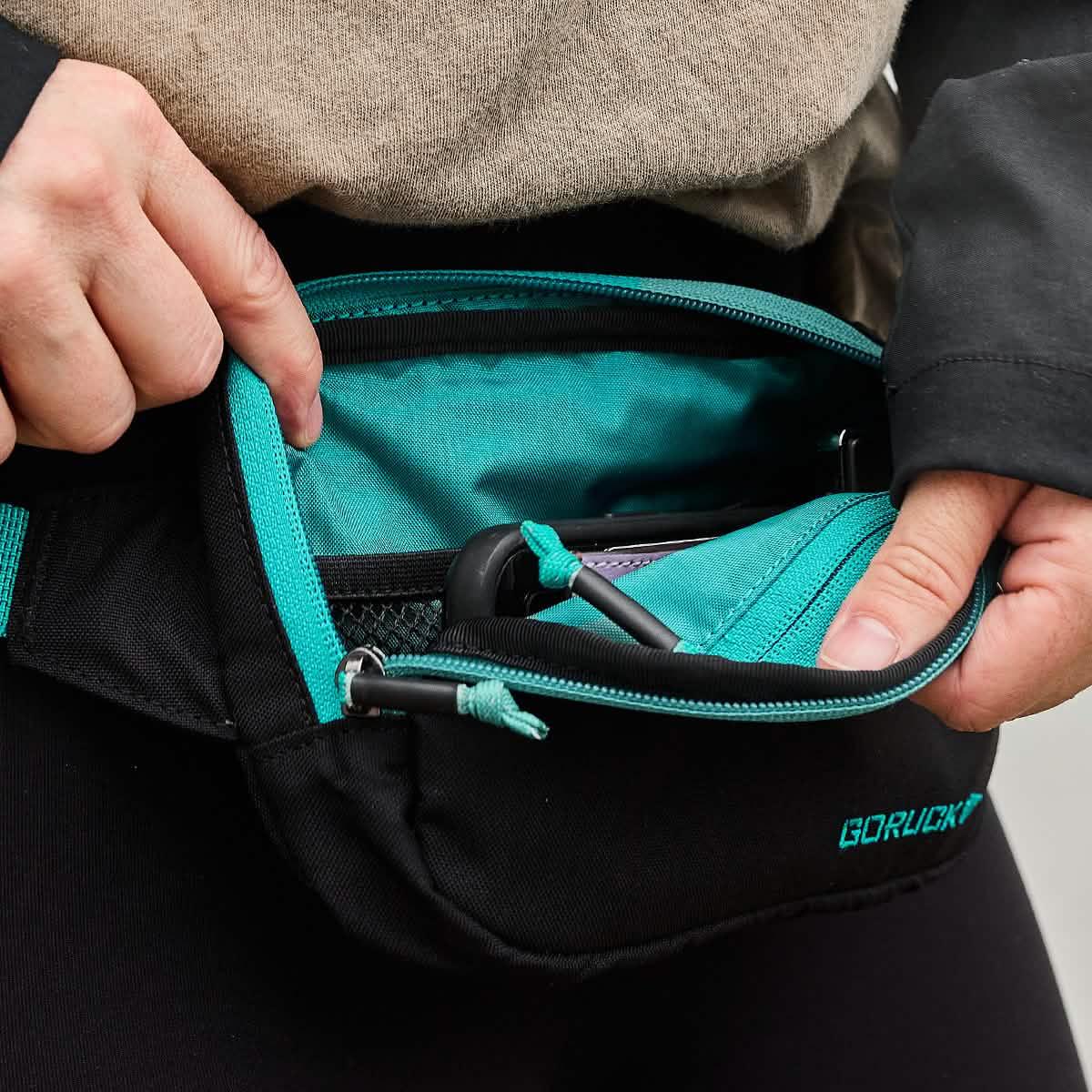 Close-up of hands opening a black and teal GORUCK belt bag showing its spacious interior