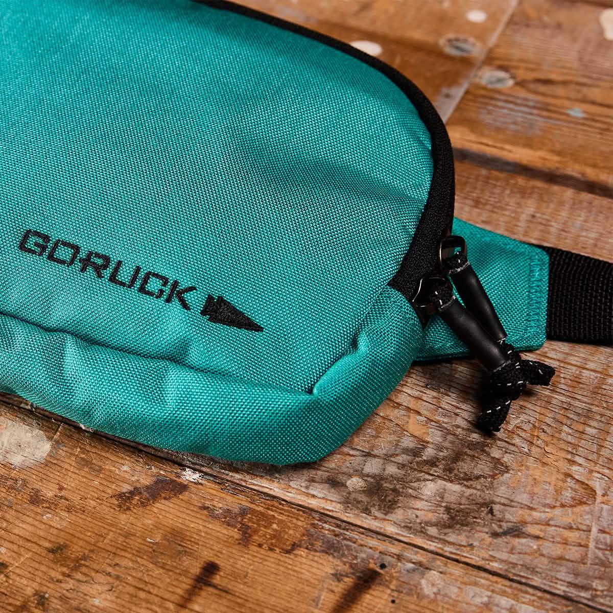 Teal GORUCK belt bag with black zipper pulls on wooden surface, rugged outdoor gear accessory