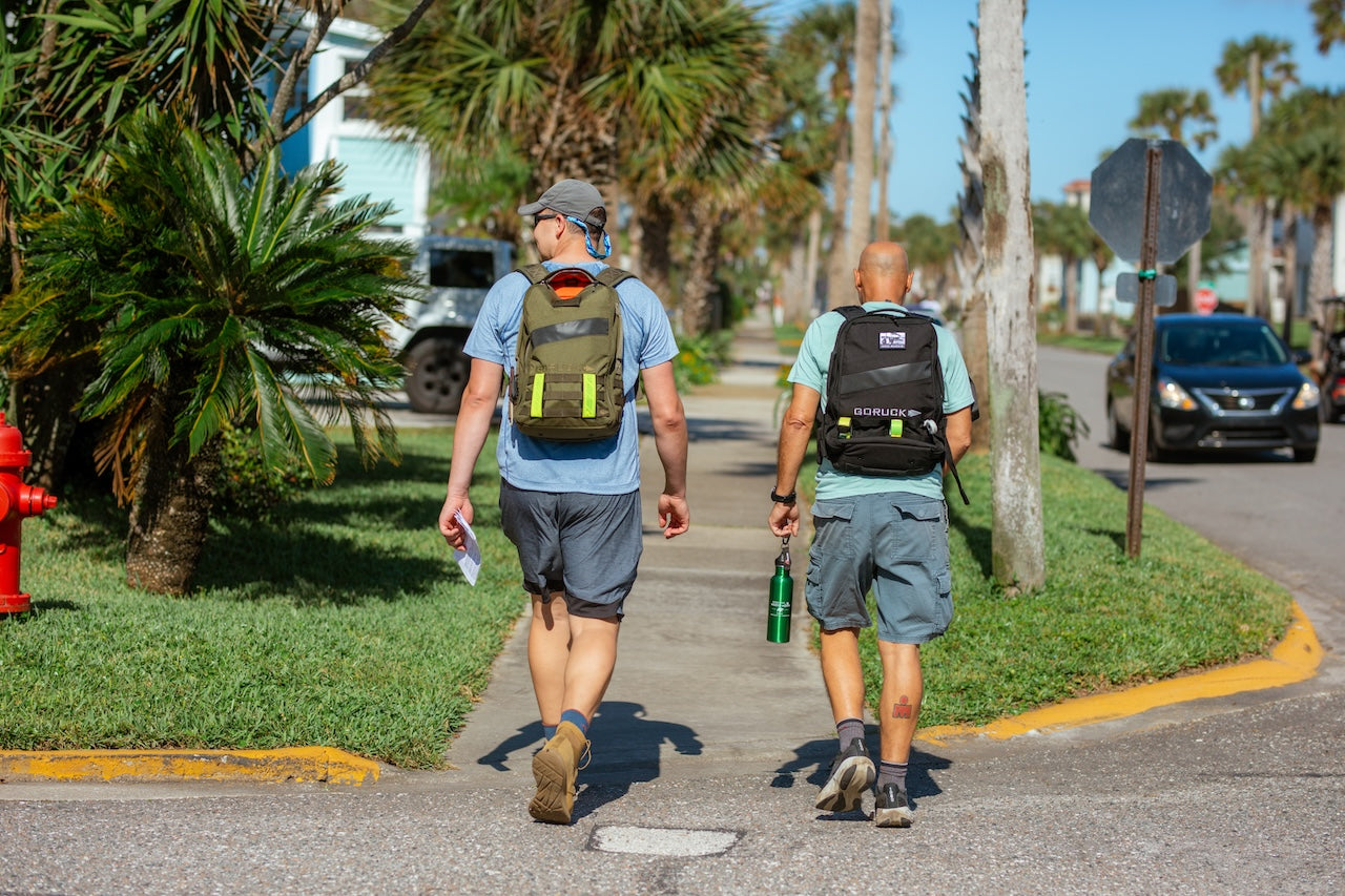 Two men with backpacks walk on a sunny sidewalk near palm trees and parked cars, looking ready for Ruckapalooza Jax Beach—Fitness Party & City Ruck Weekend.