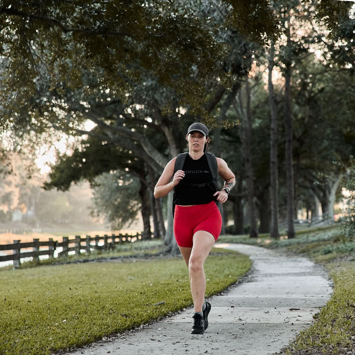 A person jogs through a scenic park, with trees swaying gently by the wooden fence, wearing GORUCK’s Women's Biker Shorts - ToughFlex for ultimate comfort and durability.