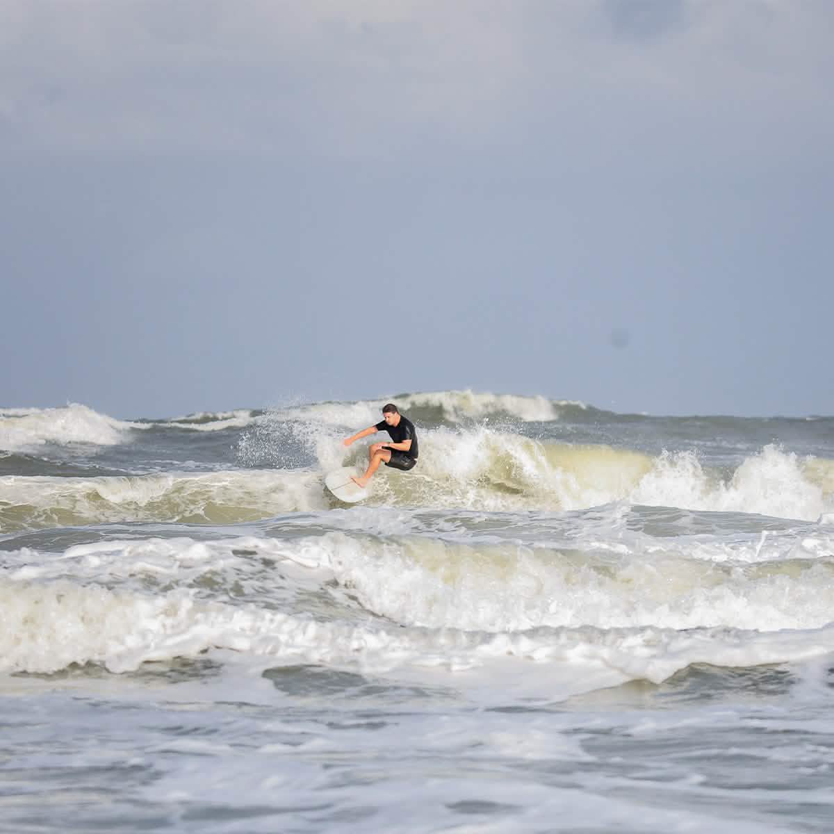 A person dressed in a black wetsuit is carving through ocean waves beneath a cloudy sky. The white, frothy swells intensify the dynamic scene as their 300 Boardshorts by GORUCK flutter in the wind.