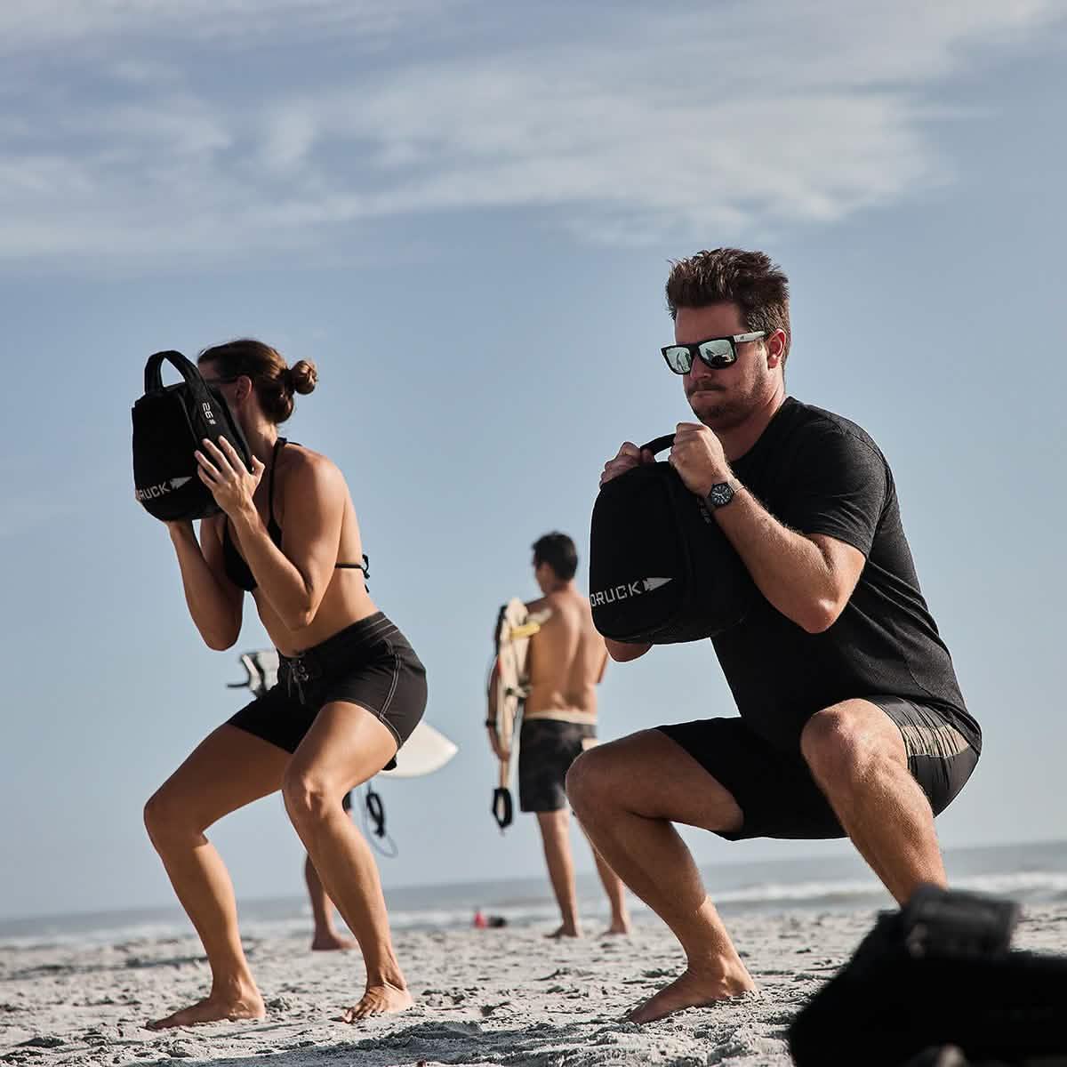 Two individuals squatting on a beach holding black GORUCK sandbags during outdoor workout