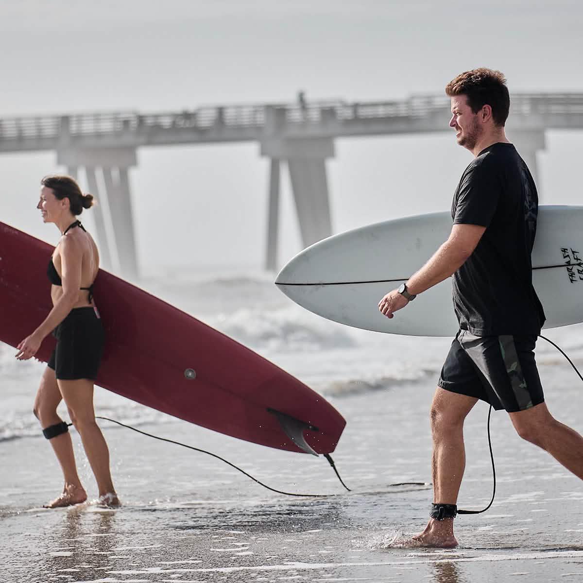 Man and woman carrying surfboards on beach near pier, both wearing black board shorts
