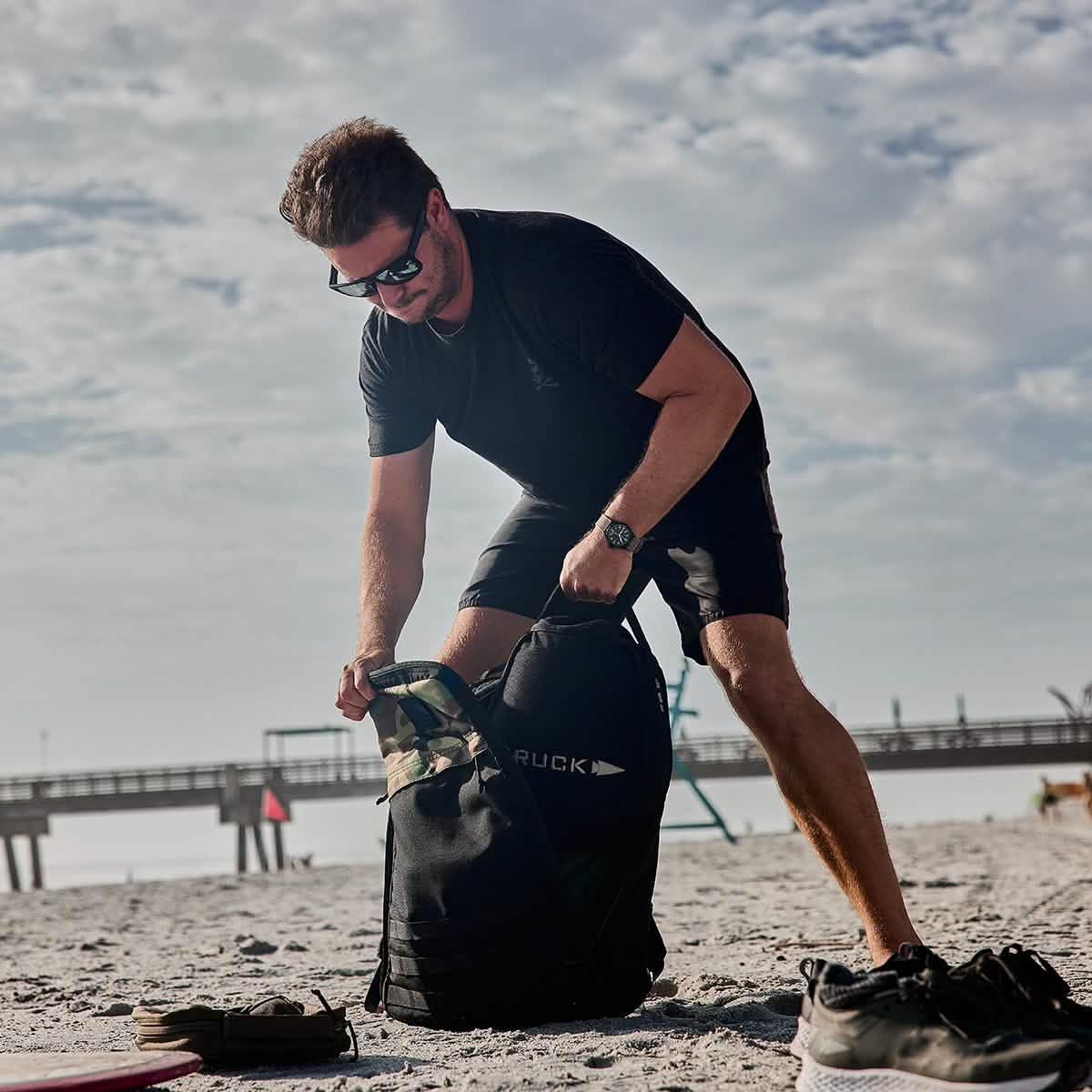 Man in black outfit packing a GORUCK backpack on a sandy beach with a boardwalk in the background